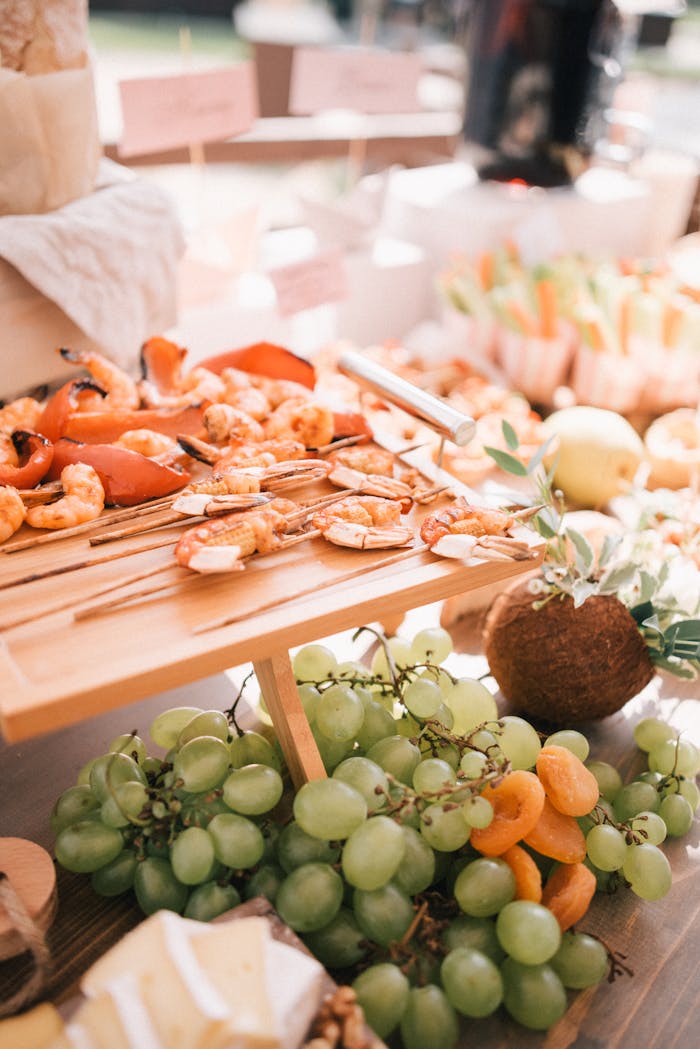 Elegant catering setup featuring skewers of shrimp, green grapes, and assorted fruits in natural lighting.